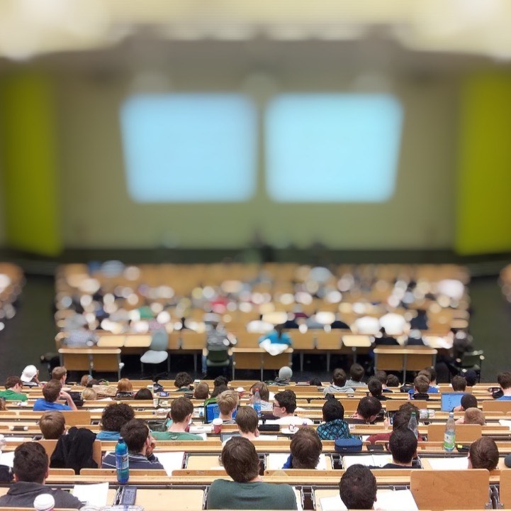 A large lecture hall filled with students at desks. The students are in focus and the front of the lecture hall with the professor and a projected presentation is out of focus.